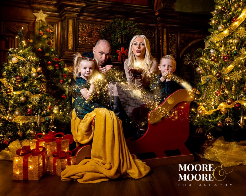 A family with their two young children enjoying a relaxed and festive traditional festive themed photographic experience in Hampshire, with lots of sparkle, Christmas Trees and Santa's sleigh. Portraits at Moore & Moore Photography