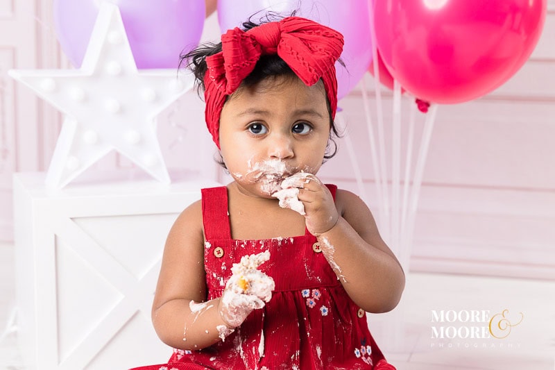 first-birthday-cake-smash-and-portraits-at-moore-moore-photography-17 One year old enjoying her cake smash cake with lots of buttercream all over her with matching colours for balloons too