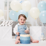 Smiling one-year-old boy enjoying a blue and white cake smash during a First Birthday Portrait Experience at Moore & Moore Photography in Fleet, Hampshire.