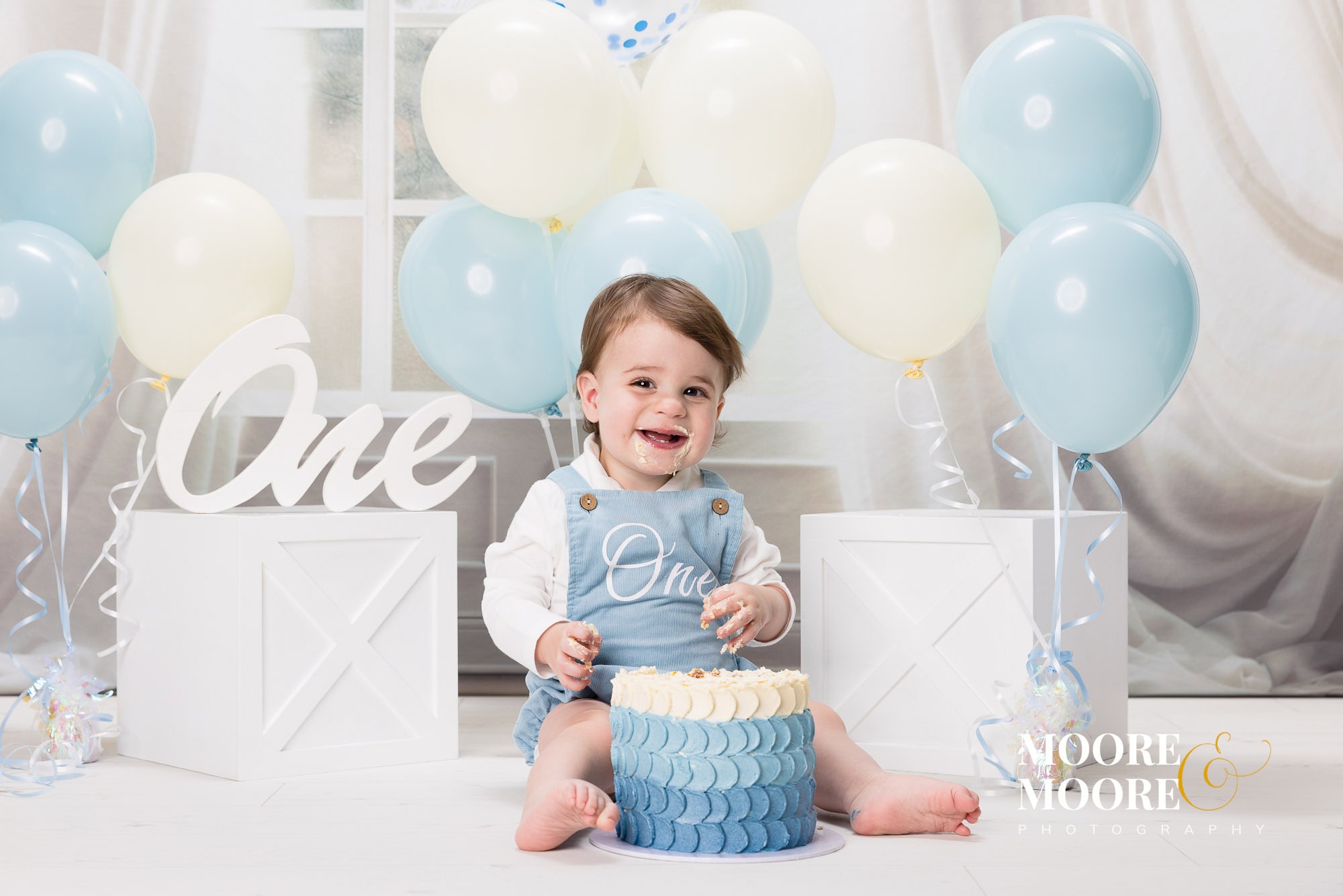First birthday cake smash portrait of a one-year-old boy surrounded by blue and cream balloons at Moore & Moore Photography studio in Fleet, Hampshire. Smiling one-year-old boy enjoying a blue and white cake smash during a First Birthday Portrait Experience at Moore & Moore Photography in Fleet, Hampshire.