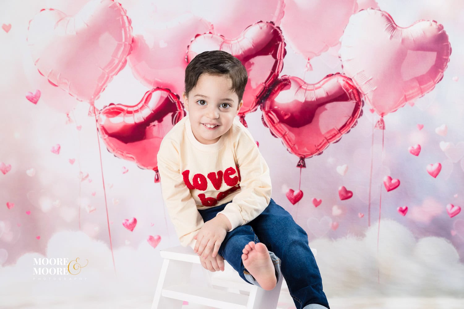 Smiling young boy sitting on a stool during a Valentine’s themed children’s portrait session at Moore & Moore Photography in Fleet, Hampshire, with pink heart balloons behind him.