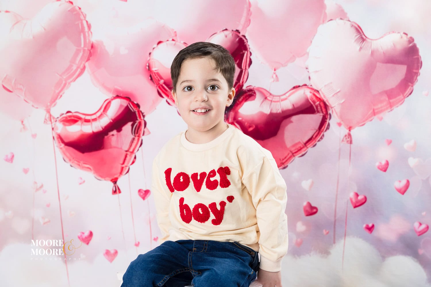 valentines-photo-experience-hampshire-13 Smiling young boy wearing a “lover boy” jumper during a Valentine’s themed children’s portrait session at Moore & Moore Photography in Fleet, Hampshire, with pink heart balloons behind him.