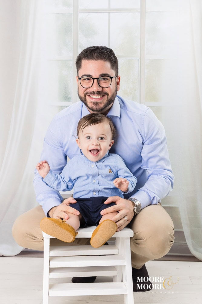 Father smiling while holding his baby during a first birthday portrait experience, showing connection and shared pride.