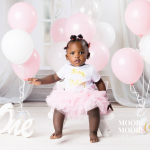One-year-old baby celebrating her first birthday, sitting confidently with soft pink and white balloons, representing joy, identity, and being deeply loved from the very beginning.
