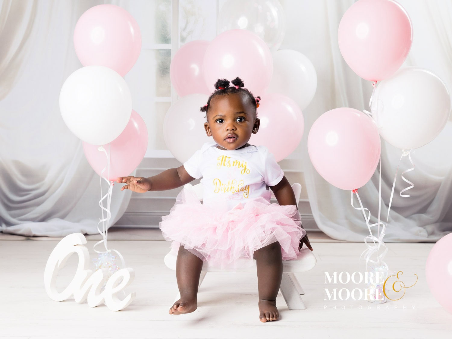 first-birthday-baby-portrait-celebrating-beginning One-year-old baby celebrating her first birthday, sitting confidently with soft pink and white balloons, representing joy, identity, and being deeply loved from the very beginning.