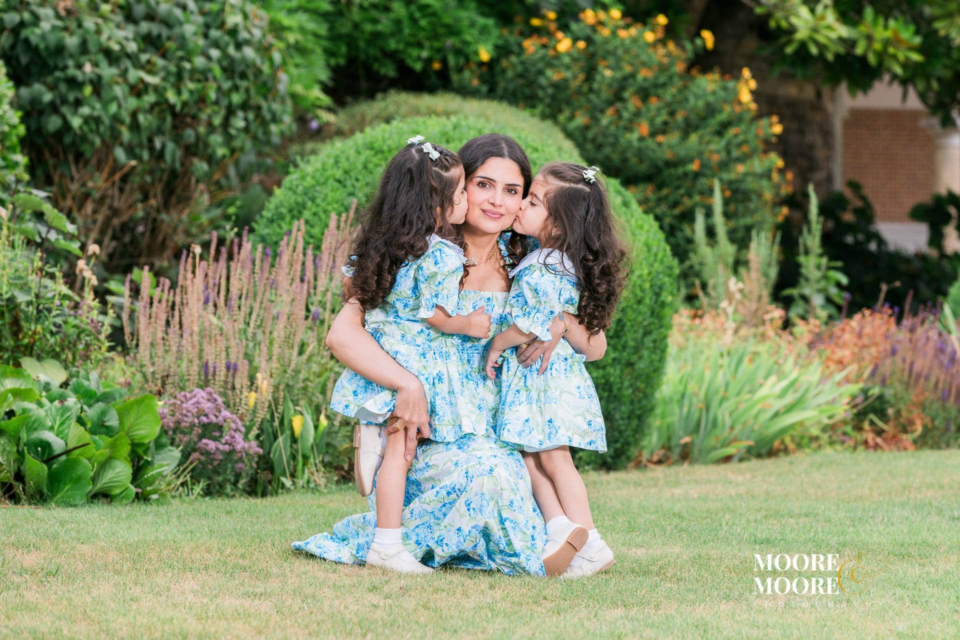 mother-daughter-affection-natural-family-portrait Two young daughters kissing their mother’s cheeks during a joyful outdoor family portrait, capturing genuine affection and connection.
