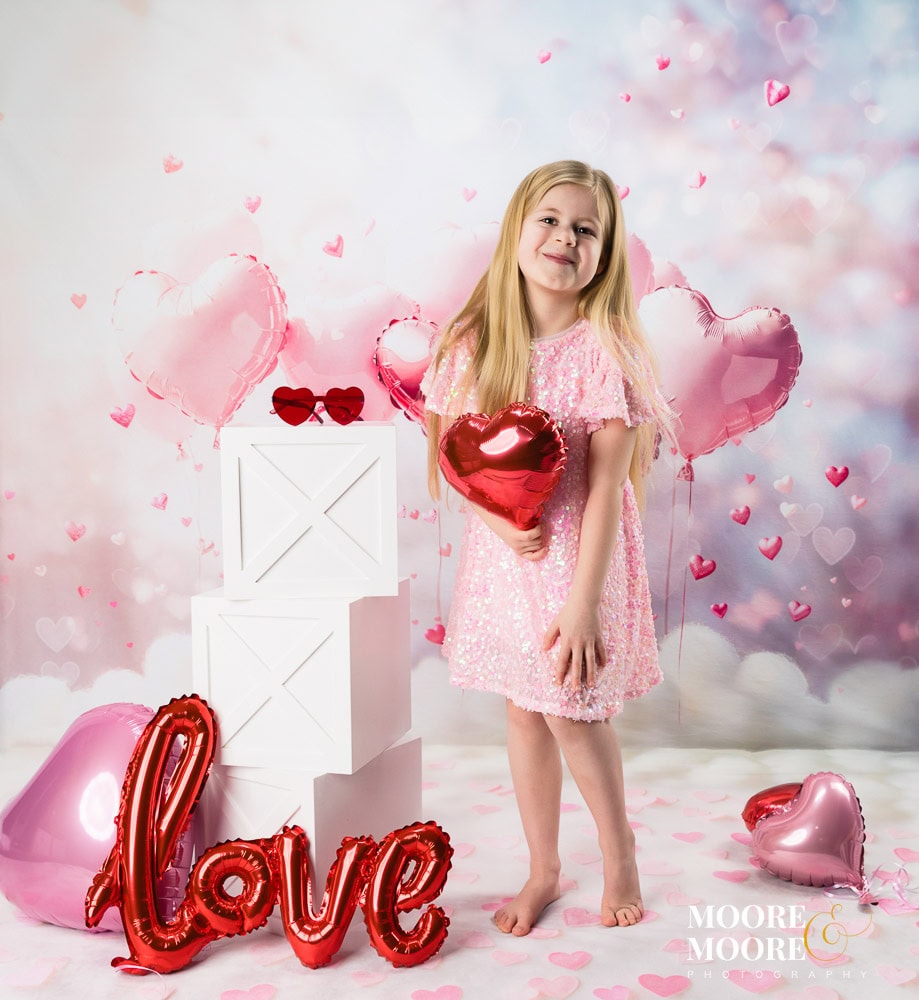 Young girl holding a heart-shaped balloon during a Valentine’s themed children’s portrait session at Moore & Moore Photography in Fleet, Hampshire, with pink balloons and “Love” letters behind her.