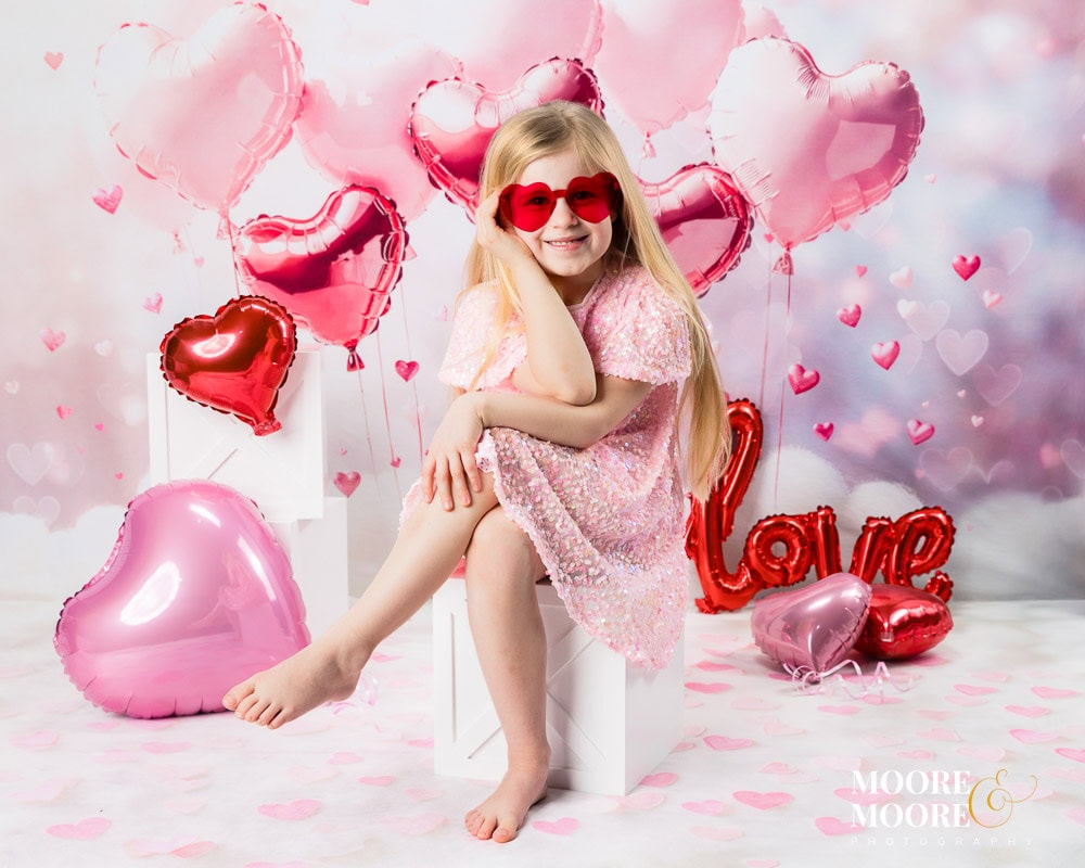 valentines-childrens-portraits-moore-and-moore-photography-fleet-hampshire Young girl wearing heart-shaped sunglasses during a Valentine’s themed children’s portrait session at Moore & Moore Photography in Fleet, Hampshire, surrounded by pink heart balloons.