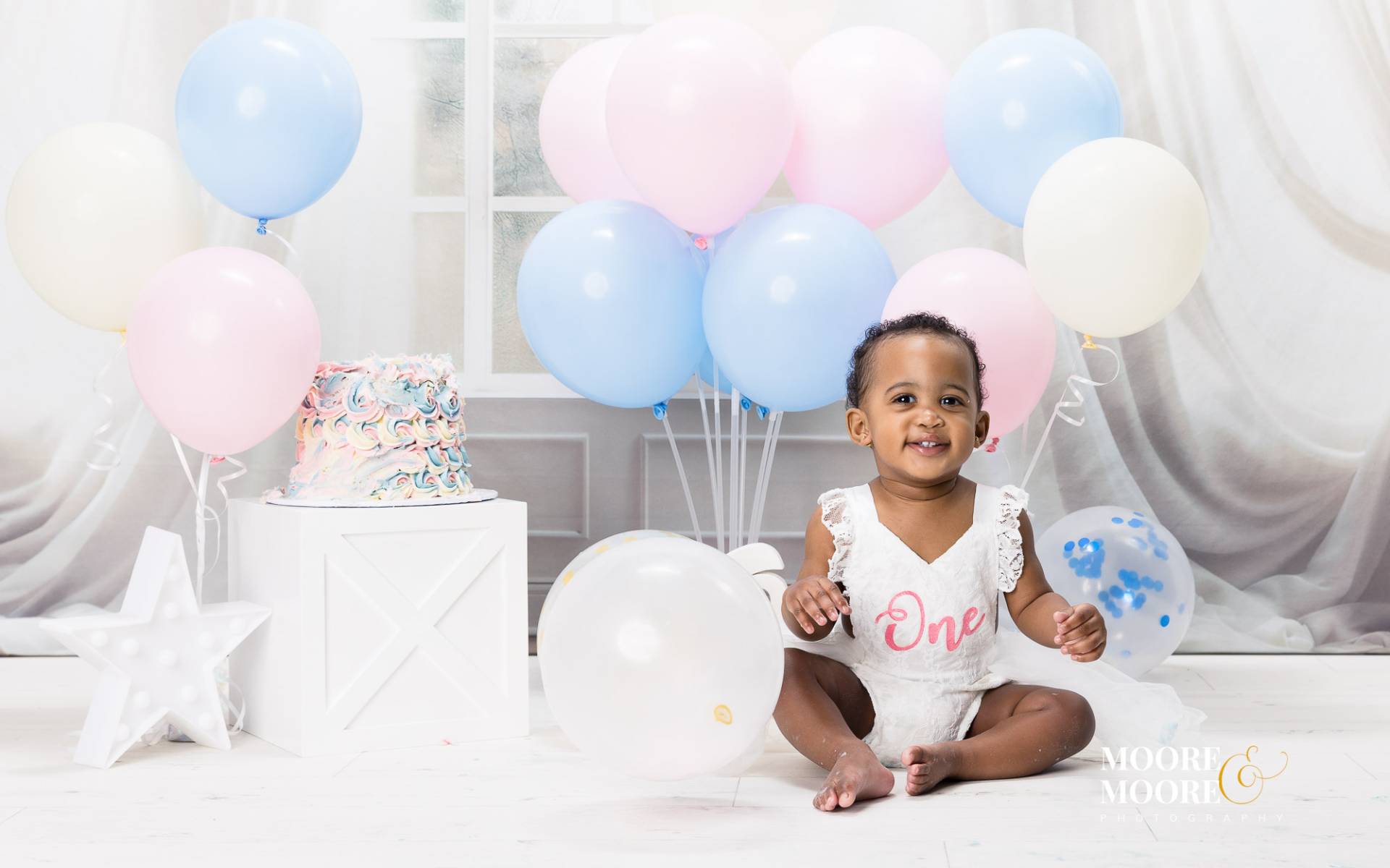 baby-first-birthday-cake-celebration-portrait smiling one year old baby sitting with birthday cake and pastel balloons during first birthday celebration