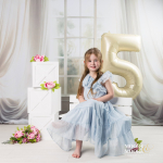 Young girl in a light blue dress sits among white decorative boxes and pink flowers, with a large gold '5' balloon behind her for a birthday portrait.