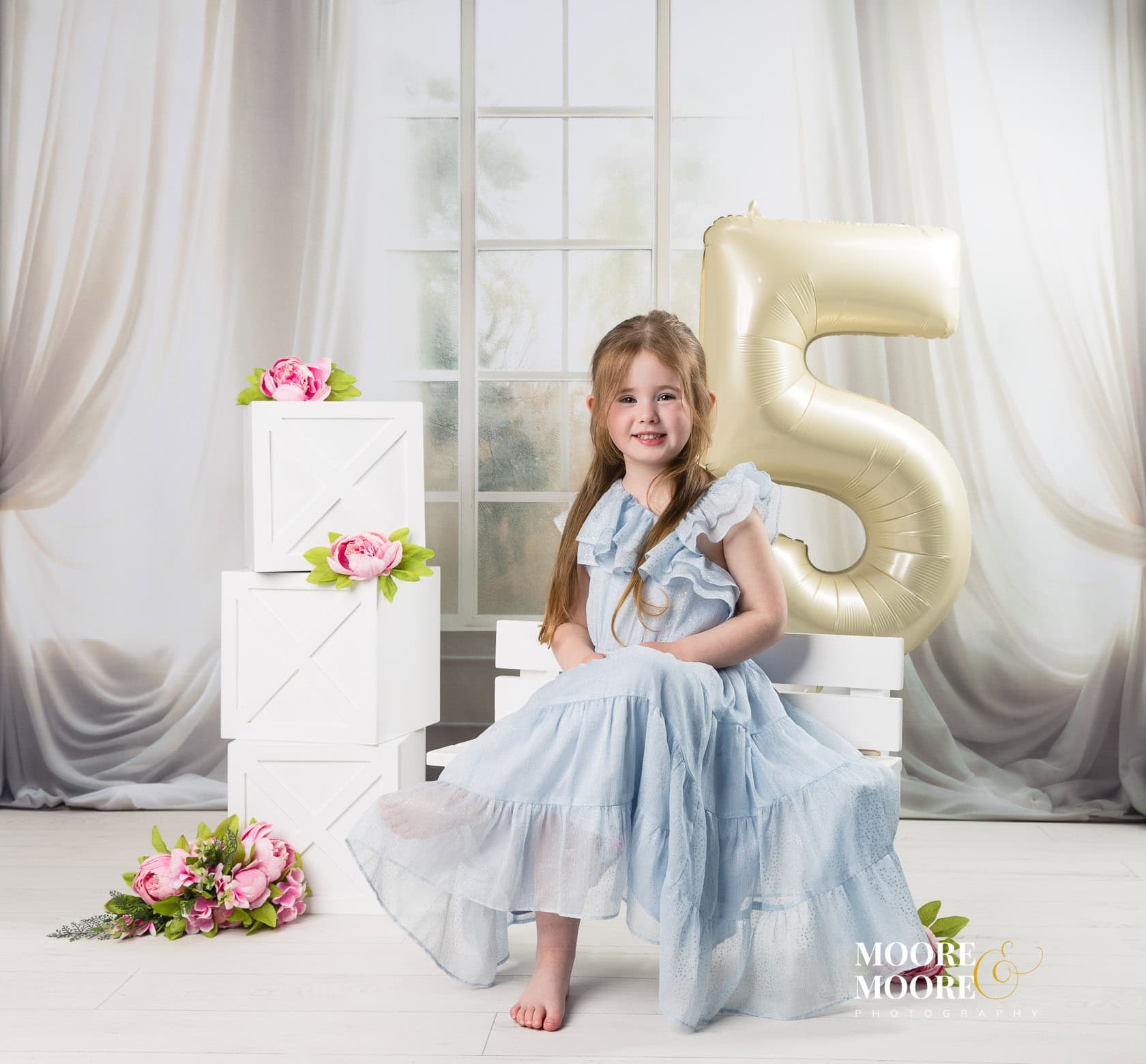 Young girl in a light blue dress sits among white decorative boxes and pink flowers, with a large gold '5' balloon behind her for a birthday portrait.